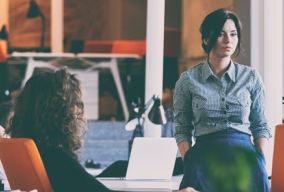 Woman sitting on desk in open plan office looking worried